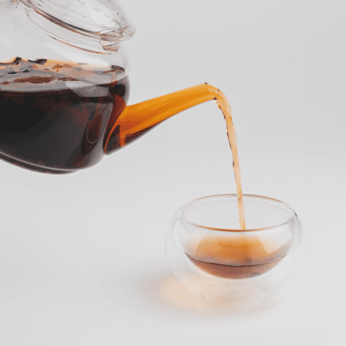 A glass teapot pouring dark, aged Puer tea into a glass cup, showcasing the deep, earthy flavors of this traditional type of tea.