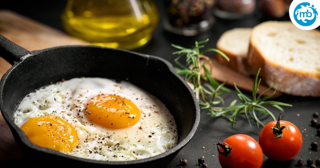 Fried eggs in a metal pan with cherry tomatoes, bread slices and olive oil kept besides the pan