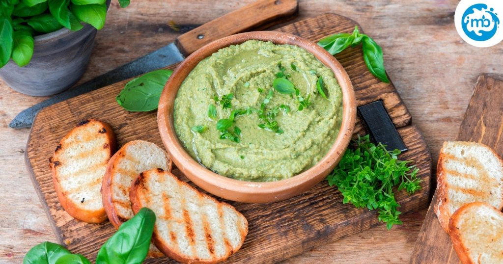 Healthy basil pesto hummus dip in a ceramic bowl, over a wooden tray, along with some toasted bread pieces.