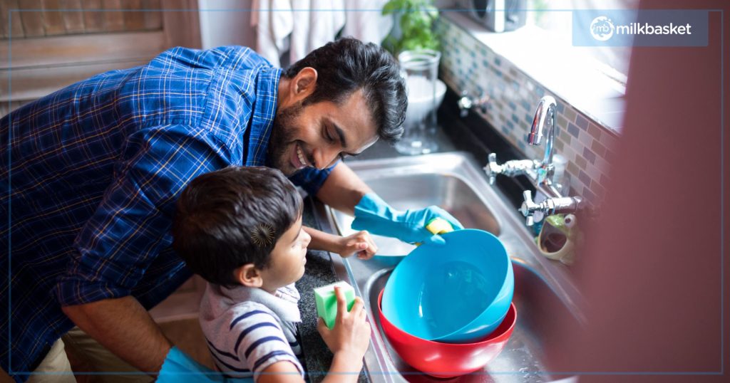 Young indian father with a child doing house chores, washing colourful dishes at the kitchen sink