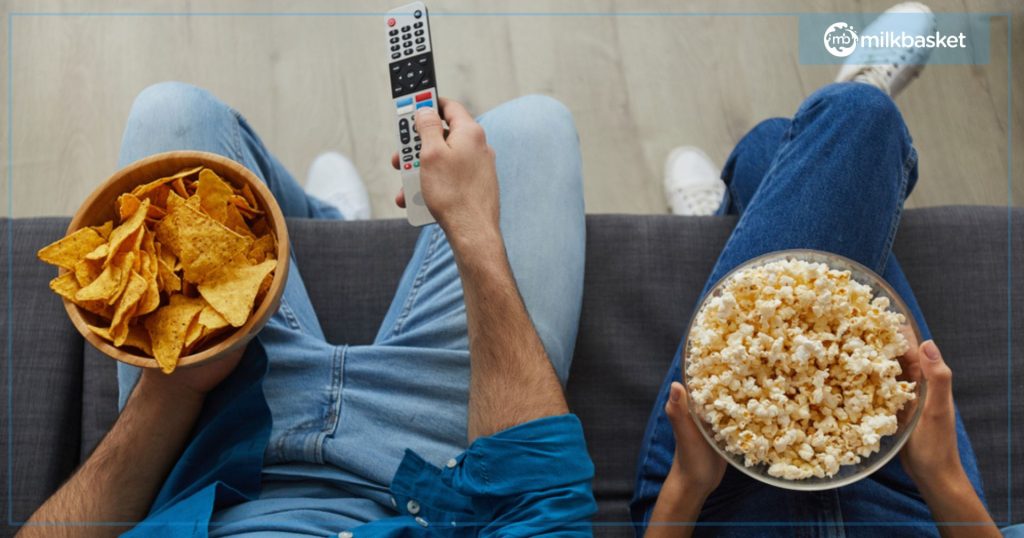 Man holding remote and a bowl of nacho chips, woman holind popcorn, watching TV on a couch