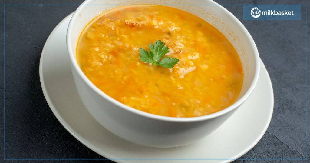 A bowl of yellow dal soup with a garnish of a single coriander leaf on top.  The bowl is white and sits on a dark stone surface