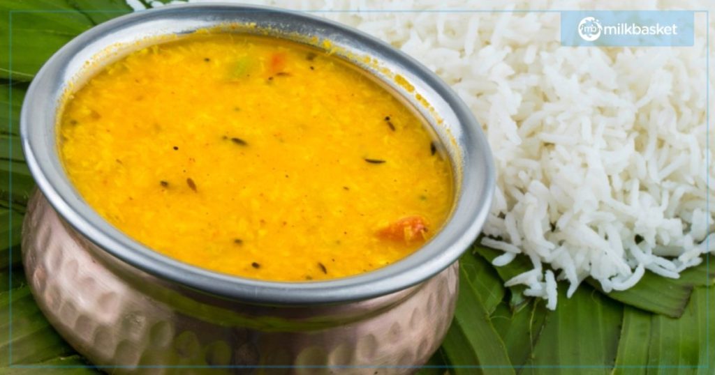 A bowl of bright, yellow Gujarati Dal served in a traditional metal dish, placed on a banana leaf with a side of fluffy white rice. The vibrant colors and rustic presentation highlight the simplicity and comfort of this classic dish.