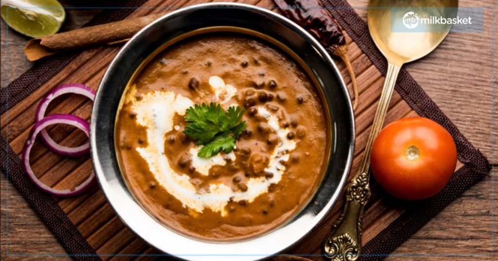 A bowl of rich, creamy Dal Makhni garnished with a swirl of cream and fresh coriander, served with sliced red onions, a tomato, and a lemon wedge on the side. A gold spoon and a dried red chili complete the rustic presentation on a wooden placemat.