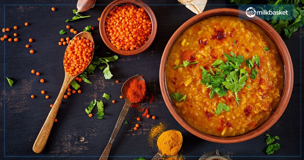 A bowl of vibrant Dal Tadka garnished with fresh coriander, surrounded by wooden spoons filled with red lentils, chili powder, and turmeric. The rustic presentation includes scattered lentils and herbs on a dark wooden surface, highlighting the rich colors and textures of the dish.