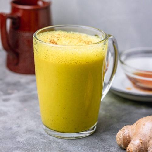 A glass mug filled with a frothy yellow beverage, likely turmeric milk, with a ginger root and a small bowl of honey visible in the background.