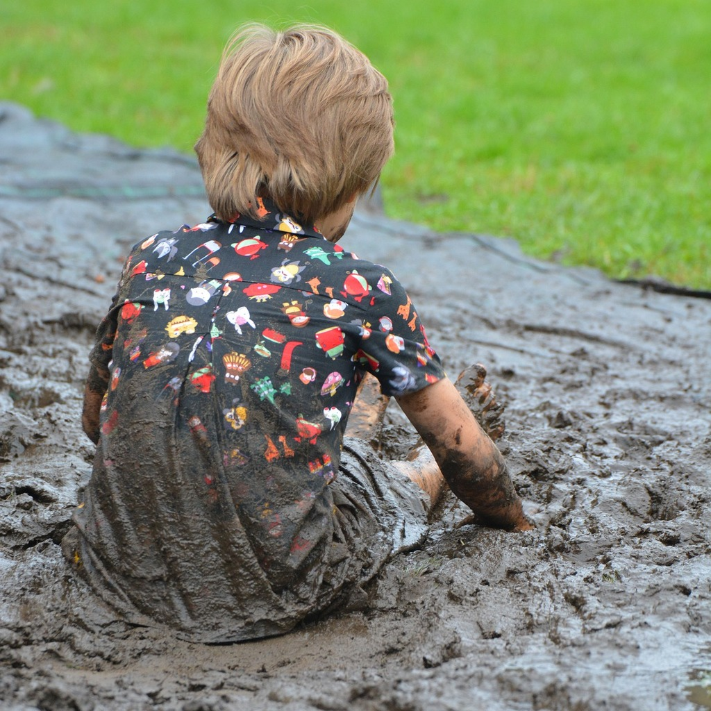 A young child with blonde hair sitting in a large muddy puddle, his clothes completely covered in mud, illustrating a common tough stain scenario.