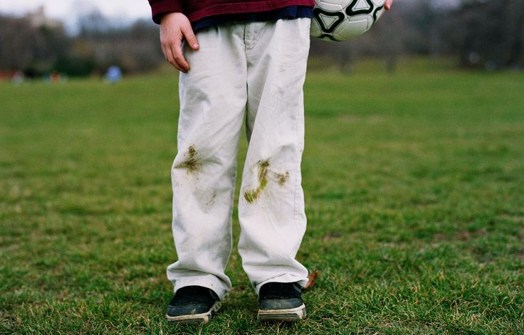 A child standing on a grassy field with visible grass stains on the knees of his white pants, holding a soccer ball, depicting stains from outdoor play.