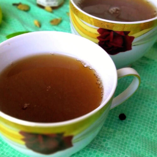 Two porcelain cups filled with a dark brown kadha,  with spices scattered around.